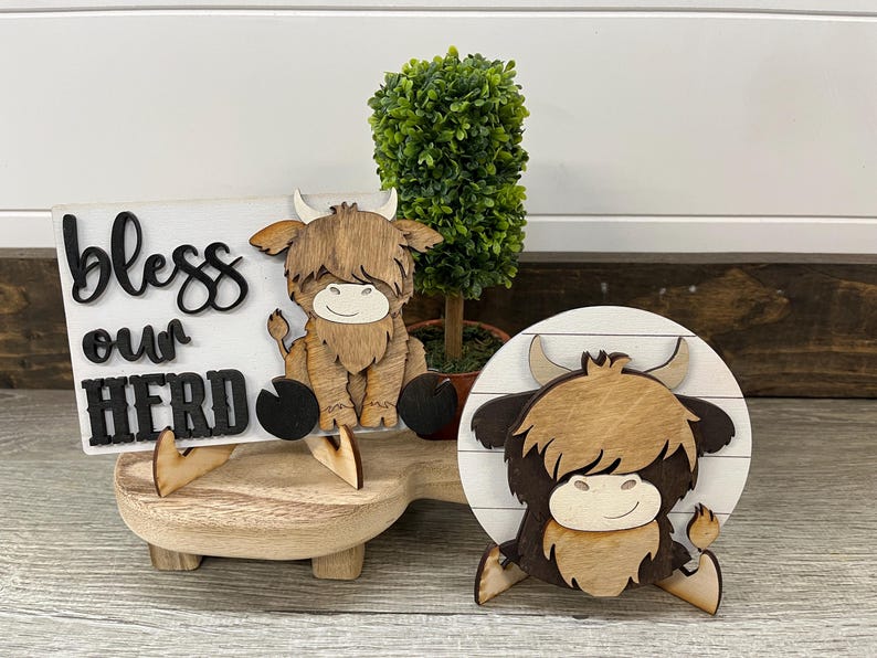 May include: Wooden farmhouse decor featuring a Highland cow and a sign that reads "bless our herd." The sign is white with black lettering. The cow figurines are brown and tan, with a smiling face. A small green plant is also included.
