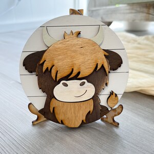 May include: A wooden Highland cow decoration. The cow is made of layered wood, featuring a light brown shaggy coat, dark brown accents, and a white and brown circular background. The cow has a smiling face and curved horns.