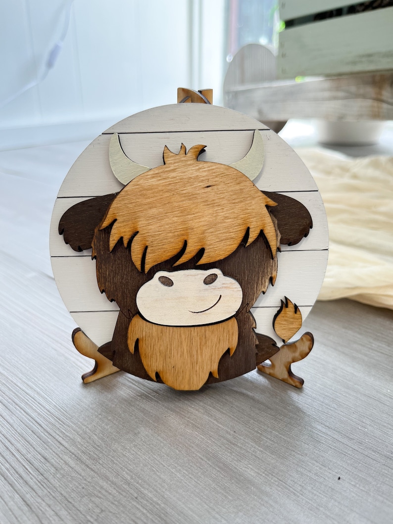 May include: A wooden Highland cow decoration. The cow is made of layered wood, featuring a light brown shaggy coat, dark brown accents, and a white and brown circular background. The cow has a smiling face and curved horns.