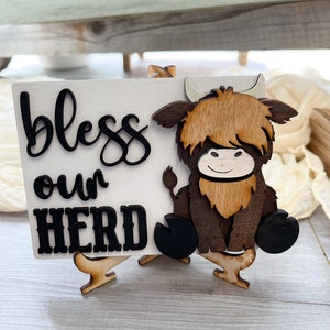 May include: A wooden sign with the words "bless our HERD" in black script and block letters. A wooden Highland cow is attached to the right side of the sign. The sign is white and sits on a small wooden easel.
