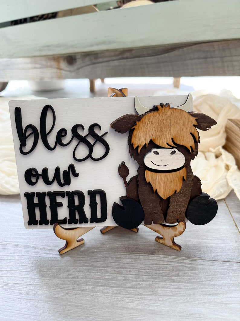 May include: A wooden sign with the words "bless our HERD" in black script and block letters. A wooden Highland cow is attached to the right side of the sign. The sign is white and sits on a small wooden easel.