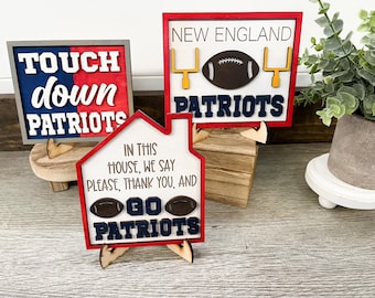 May include: Wooden signs with football-themed designs. One sign says "Touchdown Patriots" in white letters on a red and blue background. Another says "New England Patriots" with a football and goalposts. A third sign, shaped like a house, says "Go Patriots".