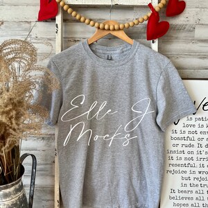 Op de afbeelding: Een grijs t-shirt met de tekst "Elle J Mocks" in wit schrift op de voorkant. Het shirt hangt aan een houten hanger met een touwtje van houten kralen en rode vilt hartjes.