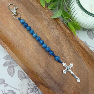 May include: A blue beaded keychain with a silver cross pendant. The keychain has round, textured blue beads strung on blue cord. A silver-coloured clasp is at the top.