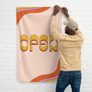 May include: A pink and orange banner with the word "open" in yellow, red, and white letters. A person is hanging the banner on a white brick wall.