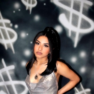 Puede incluir: Top halter de malla plateada y vaqueros blancos rotos. El fondo presenta un fondo oscuro con gráficos de signo de dólar. También es visible un collar con colgante plateado.