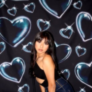 Puede incluir: Una mujer con el pelo largo y oscuro posa frente a un fondo negro con un patrón de corazones azules. Lleva un top negro corto con un detalle de corte y unos pantalones cortos de mezclilla azul oscuro. El texto "OLD SCHOOL SHOTZ" es visible en la esquina inferior derecha de la imagen.