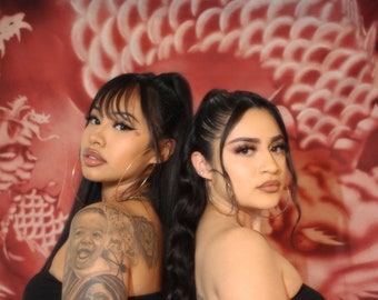 May include: Two women stand back-to-back, wearing black tube tops and blue jeans, against a red background with a white dragon design. One woman has a full arm tattoo. The image has the text "OLD SCHOOL GLAMOUR SHOTS".