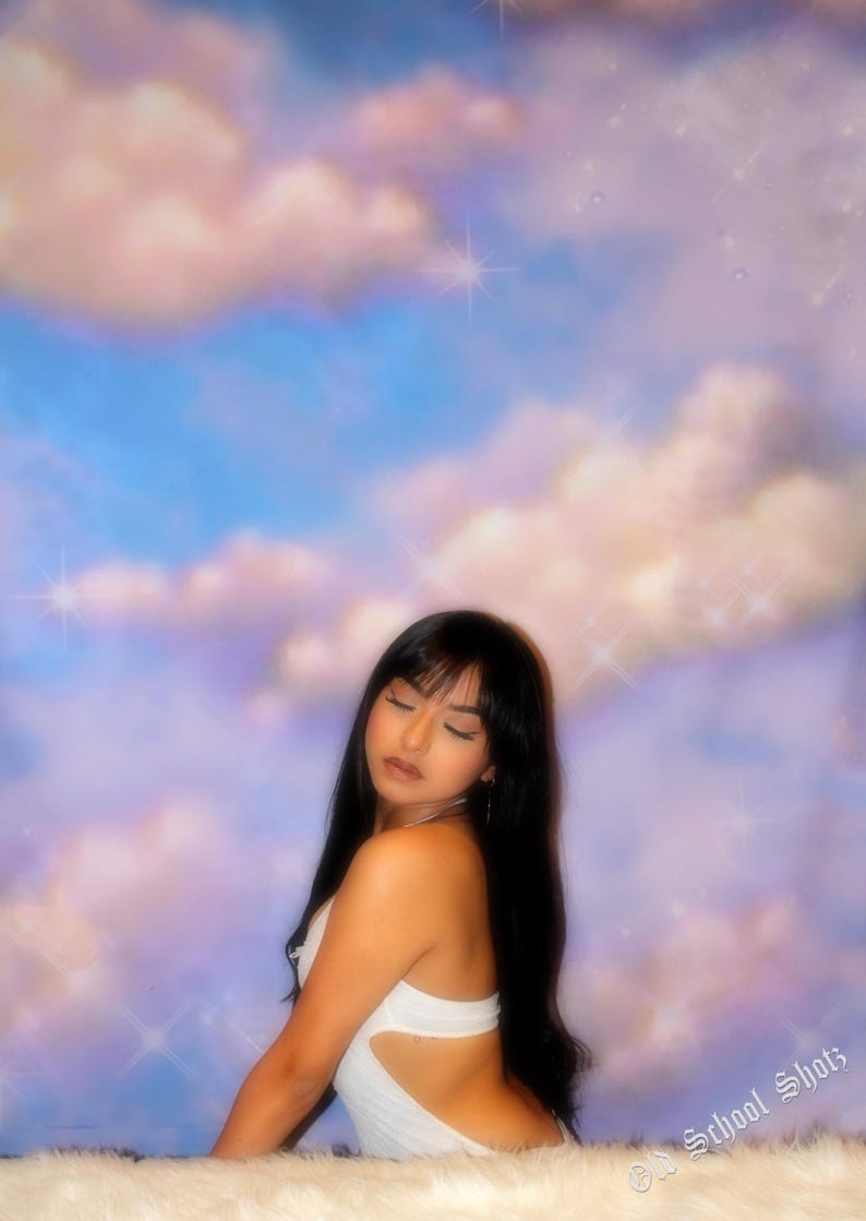 Puede incluir: Una persona con cabello largo y negro y un atuendo blanco posa contra un fondo de cielo azul con nubes rosadas y blancas esponjosas. La imagen tiene una est&eacute;tica suave y so&ntilde;adora con efectos brillantes.