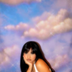 Puede incluir: Una persona con cabello largo y negro y una camiseta blanca est&aacute; acostada sobre una superficie blanca y esponjosa, con la mano apoyando la cabeza. El fondo presenta un cielo de ensue&ntilde;o con tonos azules, rosas y morados, y efectos brillantes.