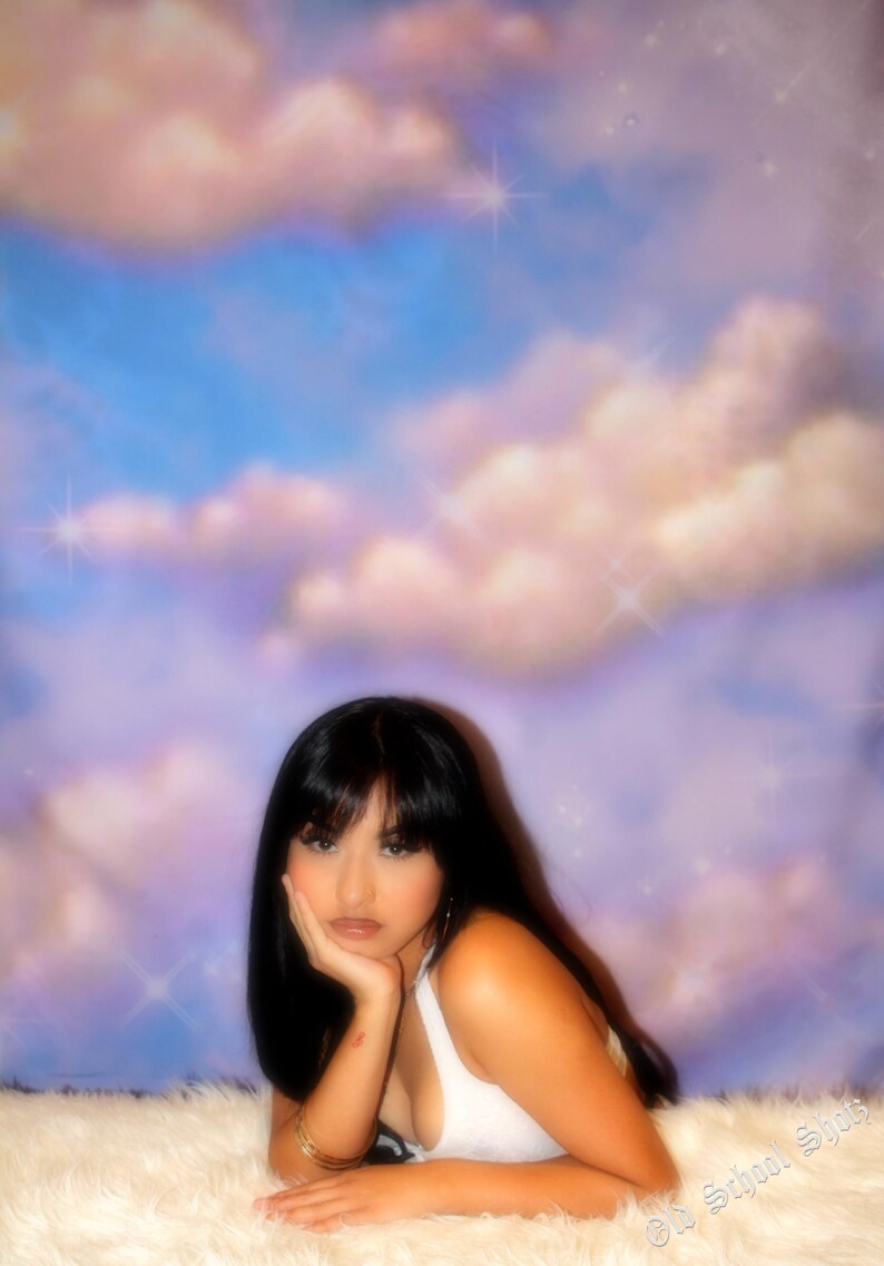 Puede incluir: Una persona con cabello largo y negro y una camiseta blanca est&aacute; acostada sobre una superficie blanca y esponjosa, con la mano apoyando la cabeza. El fondo presenta un cielo de ensue&ntilde;o con tonos azules, rosas y morados, y efectos brillantes.