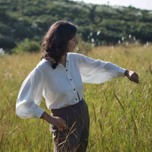 Model standing in bright green fields wearing white lotus silk shirt with one hand on thigh and the other in the air to display the fullness of the shirt's sleeve.
Background is rich green meadows and clear sky.