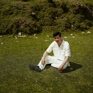 Puede incluir: Un hombre con una camisa blanca de botones y pantalones blancos se sienta en un campo de hierba. Está mirando a la cámara.
