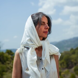Puede incluir: Una mujer con cabello gris que lleva un pa&ntilde;uelo blanco en la cabeza y una camisa blanca sin mangas con botones en la parte delantera. Est&aacute; de pie al aire libre con una colina verde en el fondo.