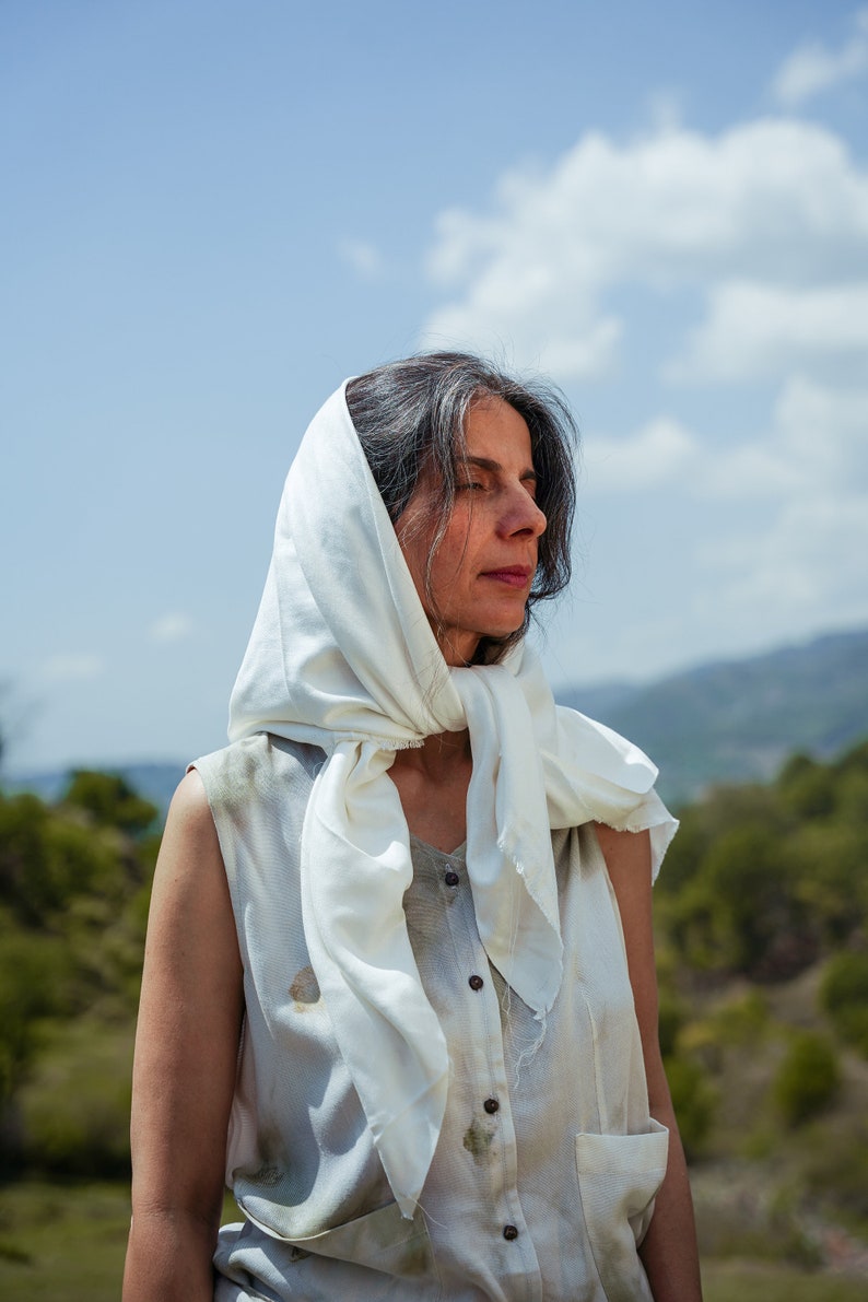 Puede incluir: Una mujer con cabello gris que lleva un pa&ntilde;uelo blanco en la cabeza y una camisa blanca sin mangas con botones en la parte delantera. Est&aacute; de pie al aire libre con una colina verde en el fondo.