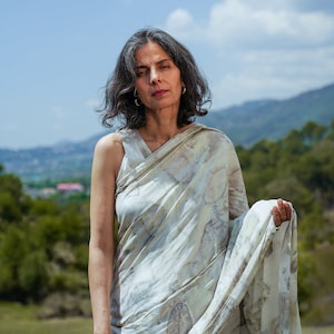 Pu&ograve; includere: Una donna indossa un sari bianco e marrone con un top senza maniche. &Egrave; in piedi in un campo con una catena montuosa sullo sfondo.
