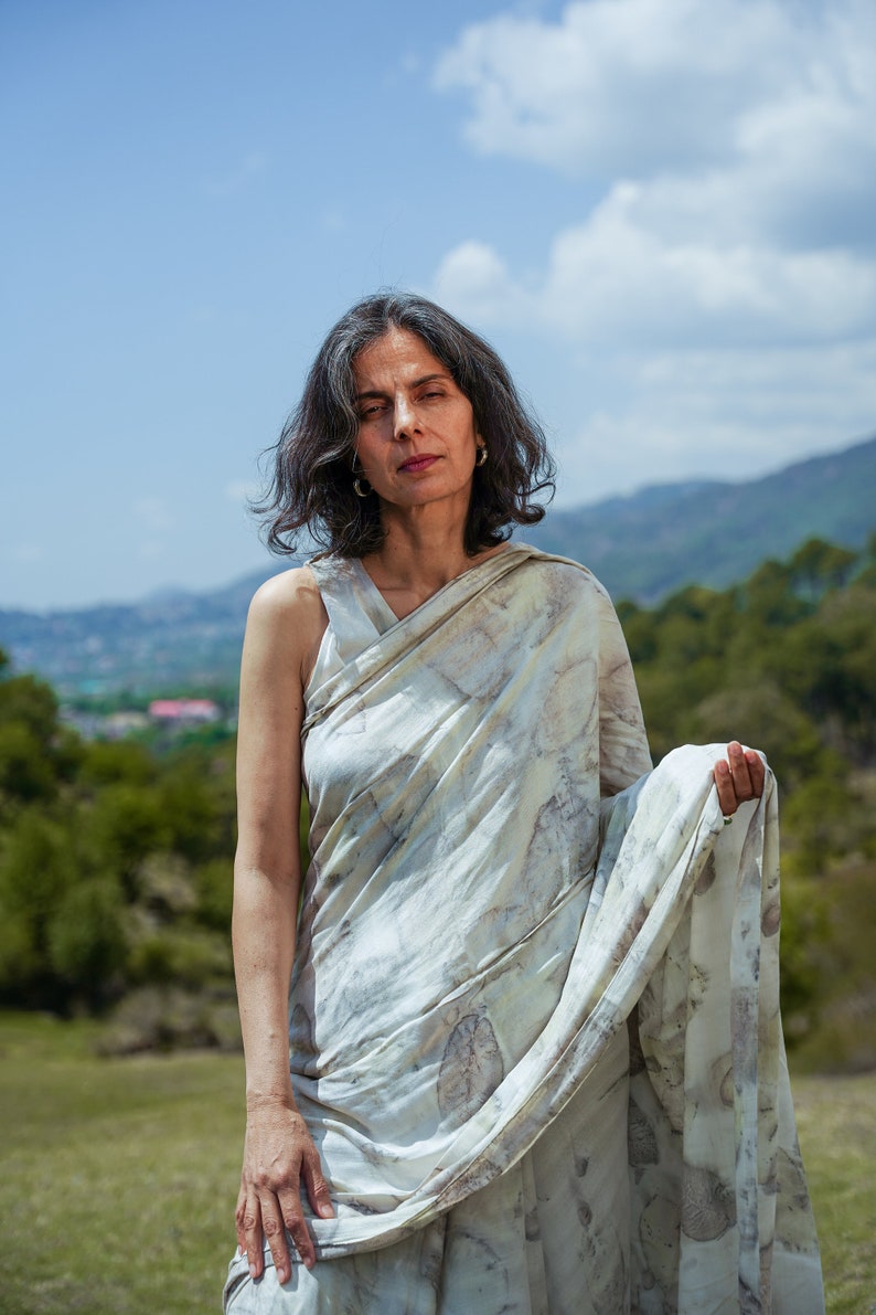 Pu&ograve; includere: Una donna indossa un sari bianco e marrone con un top senza maniche. &Egrave; in piedi in un campo con una catena montuosa sullo sfondo.