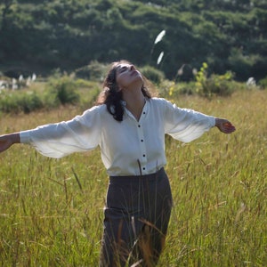 Model standing in rich green fields wearing lotus silk shirt, looking upwards with both arms open to display the fullness of the sleeves. Some tiny and blurry wildflowers in the background.