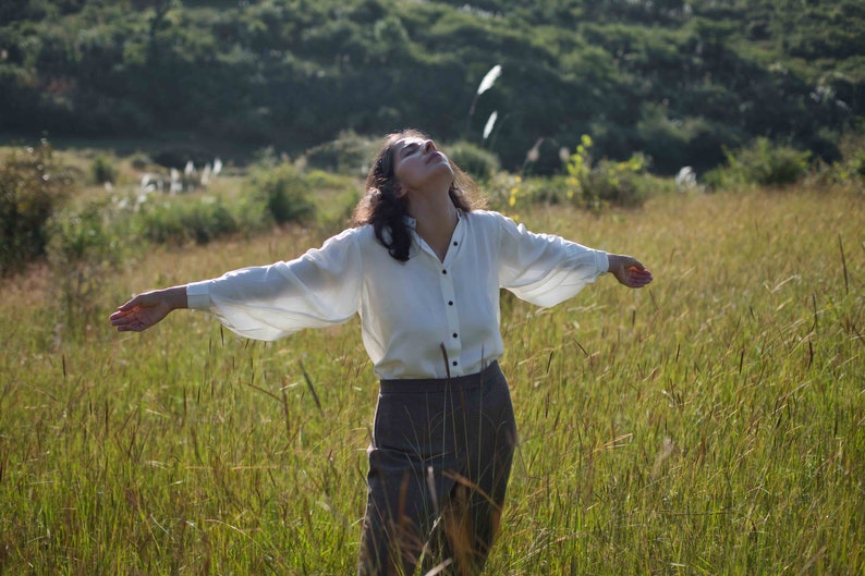 Model standing in rich green fields wearing lotus silk shirt, looking upwards with both arms open to display the fullness of the sleeves. Some tiny and blurry wildflowers in the background.