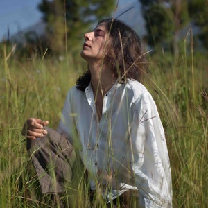 Model sitting amidst long grasses wearing white lotus silk shirt, with her hand resting on her leg, looking at the clear sky.