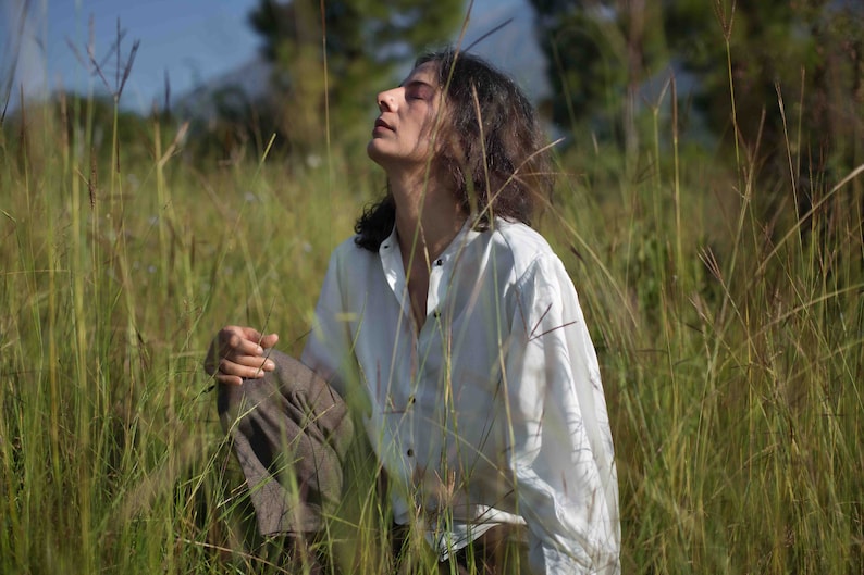 Model sitting amidst long grasses wearing white lotus silk shirt, with her hand resting on her leg, looking at the clear sky.