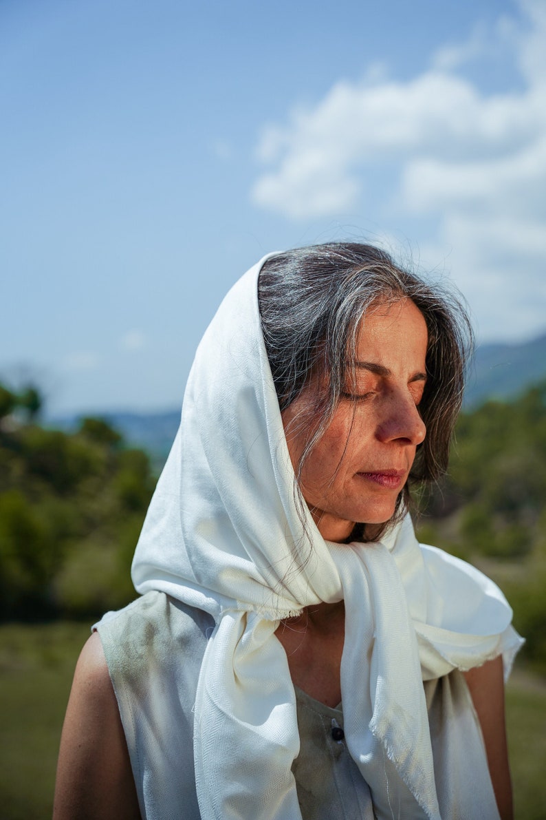 Puede incluir: Una mujer con cabello gris que lleva un pa&ntilde;uelo blanco, de pie al aire libre con un fondo verde.
