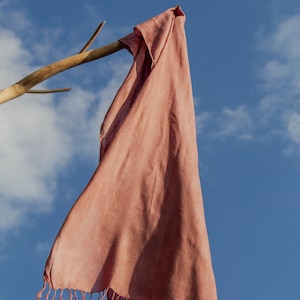 Puede incluir: Una bufanda de color rosa polvoriento con flecos colgando de una rama de madera contra un cielo azul con nubes blancas.