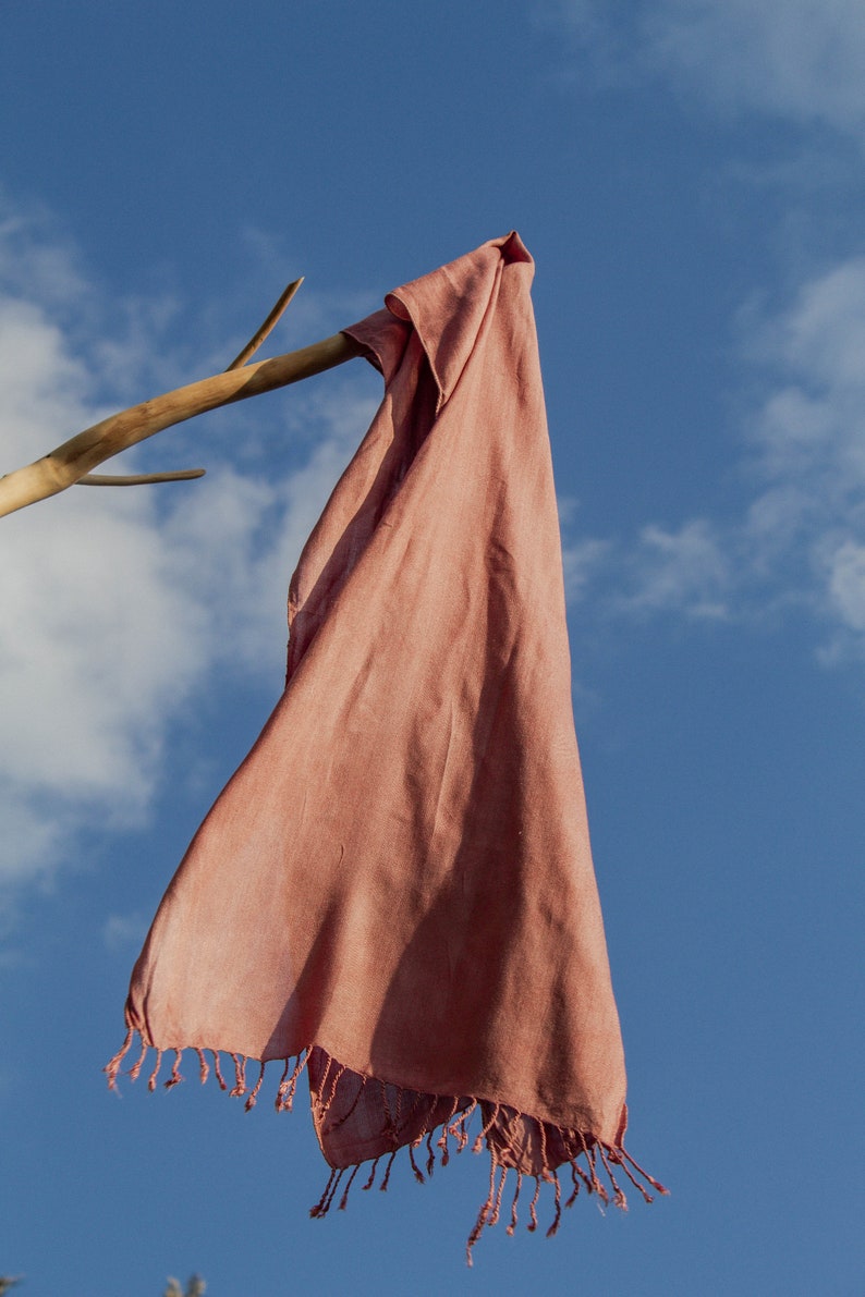 Puede incluir: Una bufanda de color rosa polvoriento con flecos colgando de una rama de madera contra un cielo azul con nubes blancas.