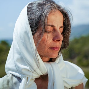Puede incluir: Una mujer con cabello gris lleva un pa&ntilde;uelo blanco en la cabeza. Tiene los ojos cerrados y mira hacia abajo. El fondo es un campo verde borroso y un cielo azul.