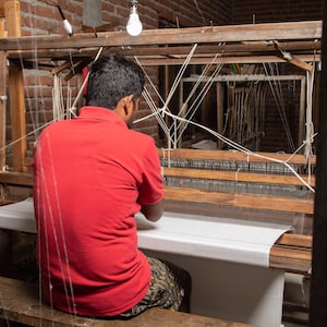 Puede incluir: Una persona con una camisa roja se sienta en un telar de madera, tejiendo una tela blanca. El telar est&aacute; hecho de madera y tiene una tela blanca estirada a trav&eacute;s de &eacute;l. La persona est&aacute; usando sus manos para tejer la tela.