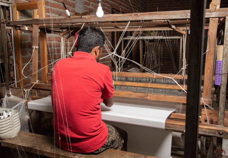 Puede incluir: Una persona con una camisa roja se sienta en un telar de madera, tejiendo una tela blanca. El telar est&aacute; hecho de madera y tiene una tela blanca estirada a trav&eacute;s de &eacute;l. La persona est&aacute; usando sus manos para tejer la tela.