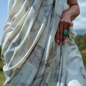 Pu&ograve; includere: Un indumento bianco e fluido con un motivo marrone chiaro e grigio. L'indumento &egrave; drappeggiato sul corpo di una persona, e un anello turchese &egrave; visibile sul suo dito.
