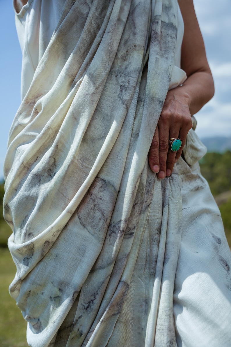 Pu&ograve; includere: Un indumento bianco e fluido con un motivo marrone chiaro e grigio. L'indumento &egrave; drappeggiato sul corpo di una persona, e un anello turchese &egrave; visibile sul suo dito.