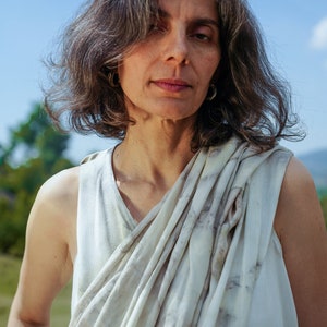 Pu&ograve; includere: Una donna con i capelli grigi indossa una canotta bianca senza maniche con una sciarpa bianca e grigia a motivi drappeggiata intorno al collo.