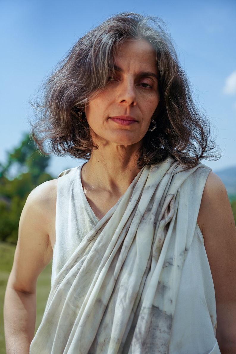 Pu&ograve; includere: Una donna con i capelli grigi indossa una canotta bianca senza maniche con una sciarpa bianca e grigia a motivi drappeggiata intorno al collo.