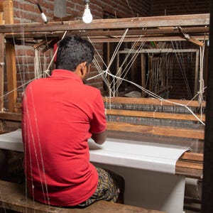 May include: A person in a red shirt sits at a wooden loom, weaving a white fabric. The loom is made of wood and has a white fabric stretched across it. The person is using their hands to weave the fabric.