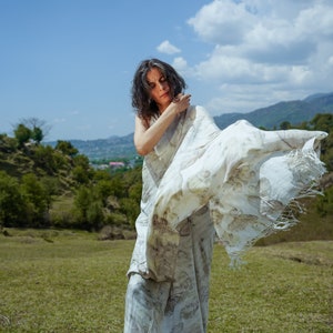Pu&ograve; includere: Una donna indossa un sari bianco e marrone con un motivo a fantasia e un tessuto fluido. &Egrave; in piedi in un campo erboso con una catena montuosa sullo sfondo.