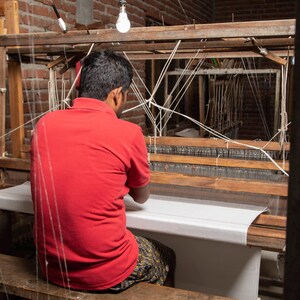 Pu&ograve; includere: Una persona con una camicia rossa &egrave; seduta a un telaio in legno, tessendo un tessuto bianco. Il telaio &egrave; in legno e ha un tessuto bianco teso su di esso. La persona sta usando le mani per tessere il tessuto.
