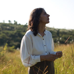 A close up of the model's upper body wearing buttoned lotus silk shirt holding wheat grass in her hands, looking slightly upwards
Background is lush green meadows, and a clear blue sky.