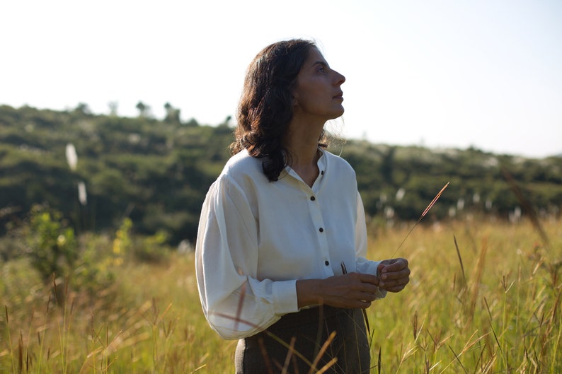 A close up of the model's upper body wearing buttoned lotus silk shirt holding wheat grass in her hands, looking slightly upwards
Background is lush green meadows, and a clear blue sky.