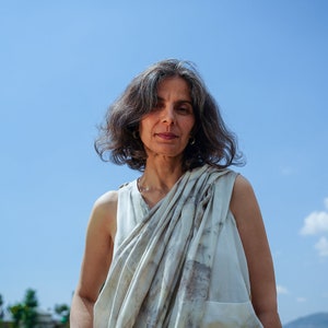 Pu&ograve; includere: Una donna con i capelli grigi indossa una canotta bianca senza maniche con una sciarpa a motivi bianchi e grigi drappeggiata sulle spalle. &Egrave; in piedi davanti a un cielo blu.
