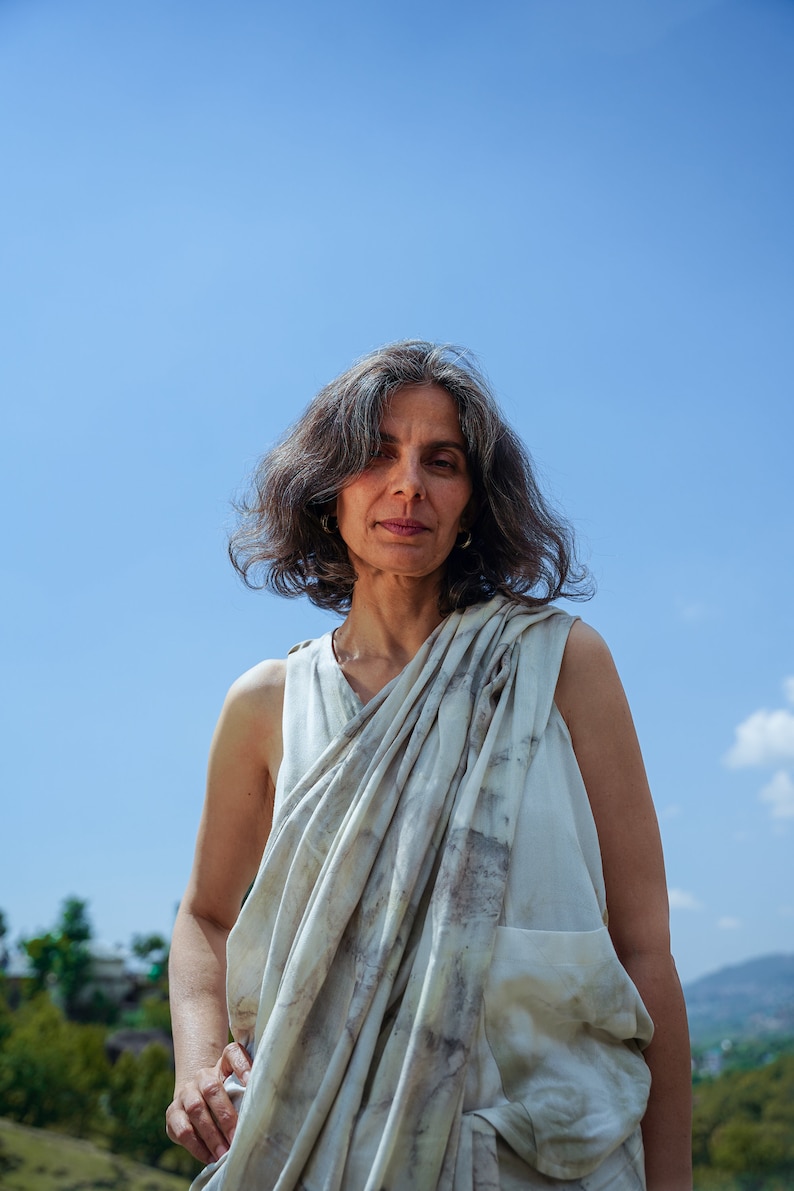 Pu&ograve; includere: Una donna con i capelli grigi indossa una canotta bianca senza maniche con una sciarpa a motivi bianchi e grigi drappeggiata sulle spalle. &Egrave; in piedi davanti a un cielo blu.