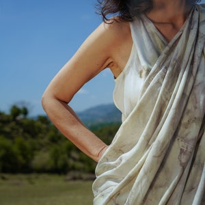 Pu&ograve; includere: Una donna indossa una canotta bianca drappeggiata senza maniche con un motivo di tintura naturale. La canotta &egrave; raccolta in vita e ha una vestibilit&agrave; ampia e fluida. La donna &egrave; in piedi in un campo con un cielo blu sullo sfondo.