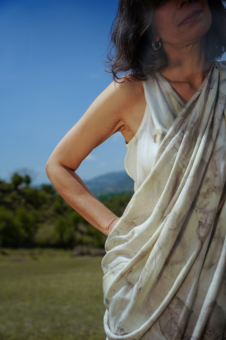 Pu&ograve; includere: Una donna indossa una canotta bianca drappeggiata senza maniche con un motivo di tintura naturale. La canotta &egrave; raccolta in vita e ha una vestibilit&agrave; ampia e fluida. La donna &egrave; in piedi in un campo con un cielo blu sullo sfondo.