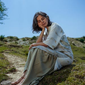 May include: A woman wearing a light beige top with a white and brown tie-dye pattern sits on a grassy hill with her head resting on her hand. She is wearing a long, light beige skirt and sandals.