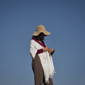 Puede incluir: Una mujer con un chal blanco con flecos, una camisa de manga larga color burdeos y un sombrero de paja de ala ancha. La mujer est&aacute; de pie contra un cielo azul brillante.