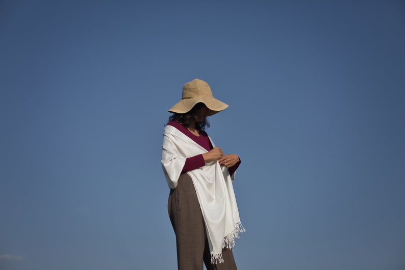 Puede incluir: Una mujer con un chal blanco con flecos, una camisa de manga larga color burdeos y un sombrero de paja de ala ancha. La mujer est&aacute; de pie contra un cielo azul brillante.