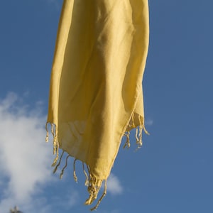 Puede incluir: Una bufanda de tela amarilla con borde de flecos cuelga contra un cielo azul con nubes blancas.