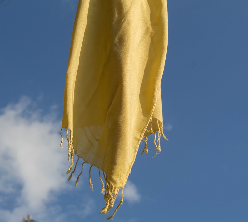 Puede incluir: Una bufanda de tela amarilla con borde de flecos cuelga contra un cielo azul con nubes blancas.