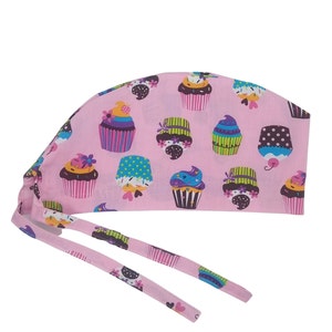 May include: Pink surgical scrub hat with a cupcake pattern. The hat has two long ties that can be tied at the back of the head.